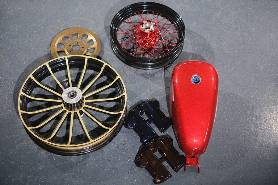 Beschichtete Motorradteile nebeneinander: Gussrad schwarz mit goldenen Speichen, Speichenrad mit roter Nabe, Riemenscheibe in Gold, roter Tank
